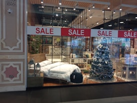 A store display showcases a cozy bedroom setup and festive decorations. A bed with white bedding is positioned beside a decorated Christmas tree with white ornaments. Surrounding furniture, including tables and chairs, adds to the homely ambiance. Red sale signs hang overhead, drawing attention to the promotional aspect.