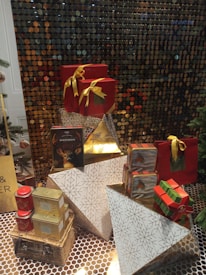 A decorative arrangement featuring several wrapped gifts in various shapes and sizes. The gifts are adorned with golden ribbons and are placed atop geometric white and golden patterned boxes. There is also a jar of shortbread with a reindeer design, a gold triangular box, and tins of other food items. The background is a shimmering, metallic sequined surface, adding to the festive vibe.