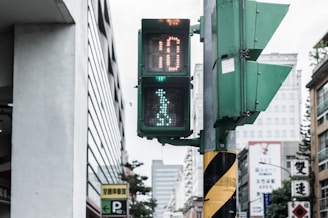 A pedestrian traffic signal displaying a walking figure within a city setting. The signal shows a countdown timer and is mounted on a pole with a yellow and black diagonal striped pattern. Surrounding the traffic light are various buildings, one with Chinese characters on signs, and a parking sign.