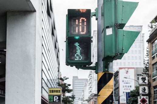 A pedestrian traffic signal displaying a walking figure within a city setting. The signal shows a countdown timer and is mounted on a pole with a yellow and black diagonal striped pattern. Surrounding the traffic light are various buildings, one with Chinese characters on signs, and a parking sign.
