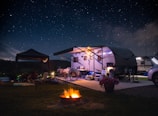 A campsite featuring a travel trailer set up under a clear, starry night sky. The scene includes outdoor furniture, bicycles, and a small tent beside the trailer. A bright, warm campfire burns in the foreground, providing a cozy atmosphere. The area is illuminated softly by lights around the trailer, and a potted plant with pink flowers adds a touch of color.