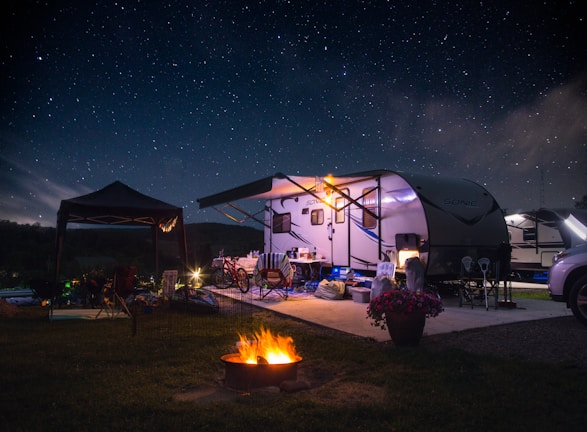 A campsite featuring a travel trailer set up under a clear, starry night sky. The scene includes outdoor furniture, bicycles, and a small tent beside the trailer. A bright, warm campfire burns in the foreground, providing a cozy atmosphere. The area is illuminated softly by lights around the trailer, and a potted plant with pink flowers adds a touch of color.