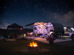 A campsite featuring a travel trailer set up under a clear, starry night sky. The scene includes outdoor furniture, bicycles, and a small tent beside the trailer. A bright, warm campfire burns in the foreground, providing a cozy atmosphere. The area is illuminated softly by lights around the trailer, and a potted plant with pink flowers adds a touch of color.