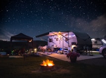 A campsite featuring a travel trailer set up under a clear, starry night sky. The scene includes outdoor furniture, bicycles, and a small tent beside the trailer. A bright, warm campfire burns in the foreground, providing a cozy atmosphere. The area is illuminated softly by lights around the trailer, and a potted plant with pink flowers adds a touch of color.
