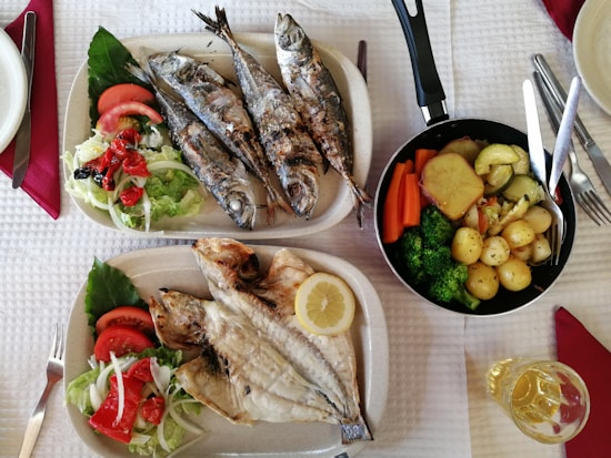 Two plates of grilled fish accompanied by fresh salad featuring lettuce, tomatoes, and red peppers, alongside a pan filled with a variety of cooked vegetables including carrots, broccoli, zucchini, and potatoes. A glass of white wine is placed nearby.