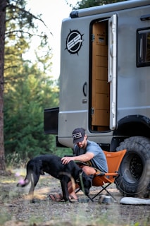 A man wearing a cap and sandals sits on an orange camping chair beside a large vehicle with off-road tires in a wooded area. He is petting a black dog that is standing next to him. The door of the vehicle is open, revealing a wooden interior.