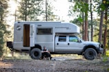A large camper truck named EarthRoamer XV-LT is parked in a forest setting. A man is sitting on the ground next to the open door of the camper, petting two dogs. The area around has tall trees and scattered sunlight.