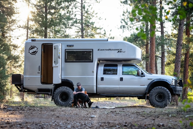 A large camper truck named EarthRoamer XV-LT is parked in a forest setting. A man is sitting on the ground next to the open door of the camper, petting two dogs. The area around has tall trees and scattered sunlight.