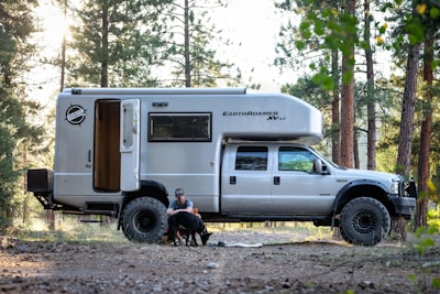 A large camper truck named EarthRoamer XV-LT is parked in a forest setting. A man is sitting on the ground next to the open door of the camper, petting two dogs. The area around has tall trees and scattered sunlight.