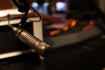 A close-up of a professional microphone is prominently featured in a recording studio setting. The background is dimly lit with blurred lights, possibly indicating an active soundboard or mixing console in use. The microphone is metallic with a mesh grille, positioned on a boom arm.