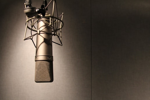 A professional microphone is suspended from a shock mount in a soundproof studio environment, with soft lighting highlighting its metallic and mesh texture against a plain backdrop.