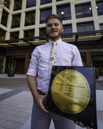 A person in a white shirt with a floral tie is holding a large square plaque with a round gold emblem. The plaque is an award for marketing and advertising, and the person is standing in front of a modern office building with large windows.