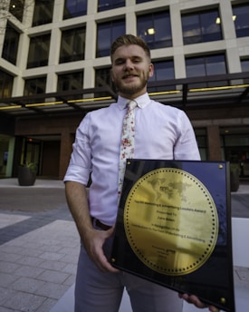 A person in a white shirt with a floral tie is holding a large square plaque with a round gold emblem. The plaque is an award for marketing and advertising, and the person is standing in front of a modern office building with large windows.