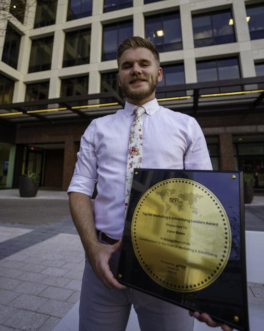 A person in a white shirt with a floral tie is holding a large square plaque with a round gold emblem. The plaque is an award for marketing and advertising, and the person is standing in front of a modern office building with large windows.