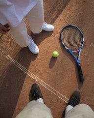 A tennis racket and ball placed on a clay tennis court, with two people standing nearby. One person is wearing white shoes and pants, while the other is wearing black shoes. The court surface is marked with white lines and the textured red-brown clay is visible.