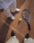 A tennis racket and ball placed on a clay tennis court, with two people standing nearby. One person is wearing white shoes and pants, while the other is wearing black shoes. The court surface is marked with white lines and the textured red-brown clay is visible.