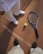 A tennis racket and ball placed on a clay tennis court, with two people standing nearby. One person is wearing white shoes and pants, while the other is wearing black shoes. The court surface is marked with white lines and the textured red-brown clay is visible.