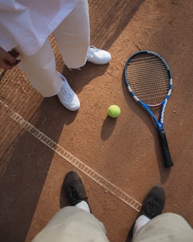 A tennis racket and ball placed on a clay tennis court, with two people standing nearby. One person is wearing white shoes and pants, while the other is wearing black shoes. The court surface is marked with white lines and the textured red-brown clay is visible.