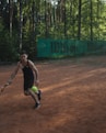 A person playing tennis on a clay court in an outdoor setting surrounded by tall trees. The person is about to hit a bright green tennis ball with a racquet and is wearing a dark sports outfit. A green banner with the word 'Wilson' is visible in the background.