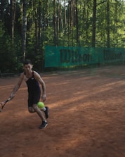 A person playing tennis on a clay court in an outdoor setting surrounded by tall trees. The person is about to hit a bright green tennis ball with a racquet and is wearing a dark sports outfit. A green banner with the word 'Wilson' is visible in the background.