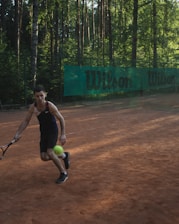A person playing tennis on a clay court in an outdoor setting surrounded by tall trees. The person is about to hit a bright green tennis ball with a racquet and is wearing a dark sports outfit. A green banner with the word 'Wilson' is visible in the background.