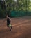 A person playing tennis on a clay court in an outdoor setting surrounded by tall trees. The person is about to hit a bright green tennis ball with a racquet and is wearing a dark sports outfit. A green banner with the word 'Wilson' is visible in the background.