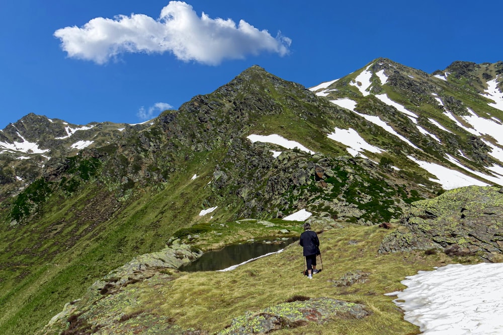 person in black jacket walking on green grass field near mountain under blue sky during daytime