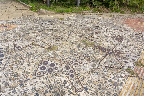 A stone mosaic embedded in the pavement featuring intricate patterns of gears and butterflies. The artwork combines various textures and shades of gray, brown, and white stones to create its design. Surrounding the mosaic are patches of grass and a small section of brick patterning.