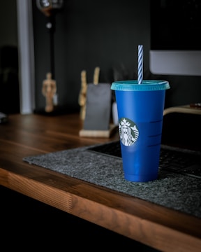 A blue Starbucks-branded reusable cup with a lid and straw sits on a dark wooden desk. The desk features a grey felt mat underneath a computer keyboard. In the blurred background, there is a small sculpture and a lamp.