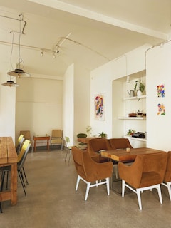 A cozy cafe interior with wooden tables and brown upholstered chairs. The walls are adorned with colorful abstract art. Pot plants are placed on shelves and tables, adding a touch of greenery. Pendant lights hang from the ceiling, providing soft lighting.