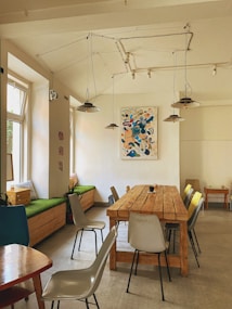 A cozy and modern interior with a long wooden table surrounded by various chairs. The space features a minimalist aesthetic with a colorful abstract painting on the wall, pendant lights hanging from the ceiling, and a bench with green cushions by the window. Natural light fills the room, adding to its inviting atmosphere.