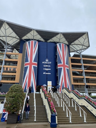 The image depicts a large, modern building with a prominent display of the Union Jack flag across its central columns. There is an extensive staircase leading up to the entrance, adorned with pink flower arrangements. Several people are present, some holding umbrellas, indicating that it might be raining.