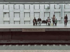 A group of people are waiting on a train platform, some sitting on benches while others stand. The setting suggests a public transport station. The wall behind the people is composed of vertical panels, and there are potted plants placed intermittently. A sign in Portuguese warns against stepping down onto the tracks.