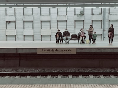 A group of people are waiting on a train platform, some sitting on benches while others stand. The setting suggests a public transport station. The wall behind the people is composed of vertical panels, and there are potted plants placed intermittently. A sign in Portuguese warns against stepping down onto the tracks.