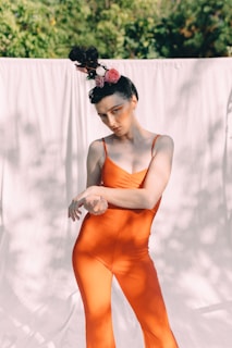 A person dressed in a bright orange jumpsuit poses outdoors. Their hair is styled in an elaborate updo adorned with pink and white flowers. The background consists of a light-colored fabric curtain with green foliage in the distance.