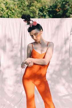 A person dressed in a bright orange jumpsuit poses outdoors. Their hair is styled in an elaborate updo adorned with pink and white flowers. The background consists of a light-colored fabric curtain with green foliage in the distance.