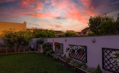A tranquil backyard garden with a lush green lawn is bordered by a low wall adorned with trellises and hanging flower pots. Vibrant flowers add color to the scene. Trees and bushes line the edge, while houses in the background are silhouetted against a stunning sunset sky filled with pink and orange clouds.