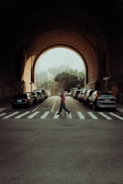 A person wearing casual clothing is walking across a zebra crossing under an archway. The background features parked cars aligning both sides of the street, and soft sunlight is filtering in, casting shadows and creating a warm, moody atmosphere.