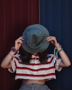 A person wearing a striped crop top and holding a hat over their face, standing in front of a dual-colored background, half red and half blue. The person is adorned with multiple bracelets and rings, creating a casual and stylish appearance.