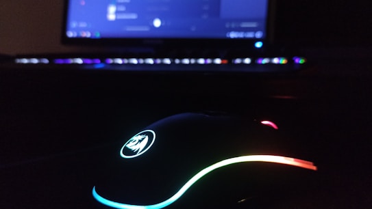 A gaming setup with a focus on a colorful RGB-lit mouse in the foreground, and a softly glowing keyboard and monitor in the background. The surroundings are dimly lit, enhancing the contrast and the vivid colors of the lights.