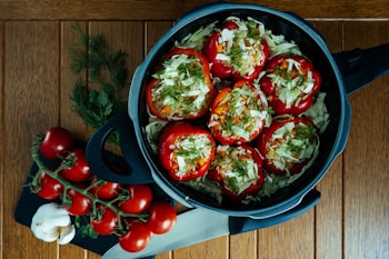 A dark pot sits on a wooden table, filled with red bell peppers stuffed with chopped cabbage, rice, and herbs. Fresh tomatoes on the vine, garlic cloves, and sprigs of fresh herbs lie on a cutting board beside the pot. A kitchen knife is placed nearby, completing the preparation scene.