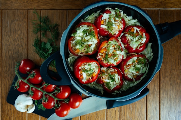 A dark pot sits on a wooden table, filled with red bell peppers stuffed with chopped cabbage, rice, and herbs. Fresh tomatoes on the vine, garlic cloves, and sprigs of fresh herbs lie on a cutting board beside the pot. A kitchen knife is placed nearby, completing the preparation scene.