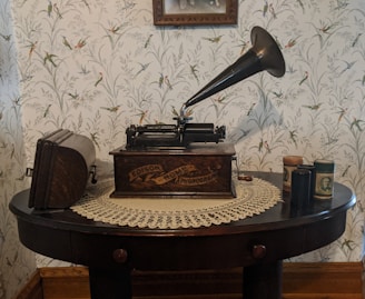 A vintage Edison phonograph sits atop a dark wooden table covered with a lace doily. The phonograph's large horn and crank are prominent, and there are several cylindrical containers on the table, likely phonograph cylinders. The background features floral wallpaper with bird motifs, adding to the antique ambiance.