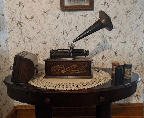 A vintage Edison phonograph sits atop a dark wooden table covered with a lace doily. The phonograph's large horn and crank are prominent, and there are several cylindrical containers on the table, likely phonograph cylinders. The background features floral wallpaper with bird motifs, adding to the antique ambiance.