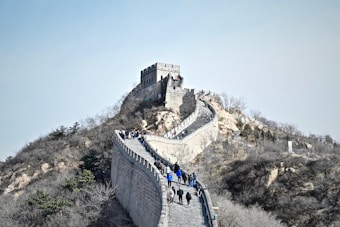 A section of the Great Wall of China stretches across a rugged hillside, with tourists walking along the stone pathway. The ancient fortification features a watchtower at the highest point, surrounded by bare trees and rocky terrain under a clear sky.