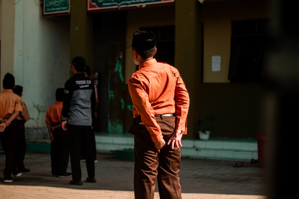 オレンジ色の長袖シャツと茶色のズボンを着た男性が、歩道を歩いている人の近くに立っています。