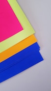 Four colorful sheets of paper are layered, with the top sheet being fluorescent pink, followed by a bright yellow, orange, and blue sheet. They are arranged neatly on a light gray surface.