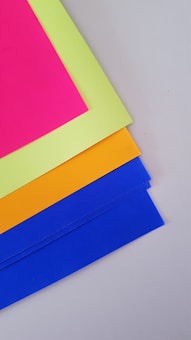 Four colorful sheets of paper are layered, with the top sheet being fluorescent pink, followed by a bright yellow, orange, and blue sheet. They are arranged neatly on a light gray surface.