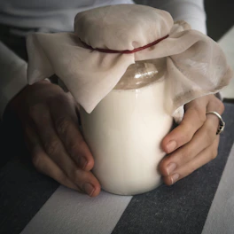 Hands gently holding a small jar of creamy, natural ferment surrounded by terracotta and linen.