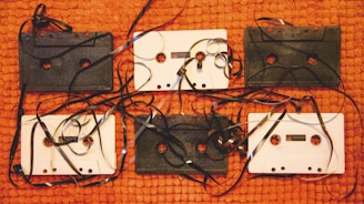 Several vintage audio cassette tapes are displayed on a textured orange surface. The tapes are intermingled with unwound, tangled magnetic tape, creating a chaotic yet nostalgic arrangement. The contrast between black and white cassettes adds visual interest.